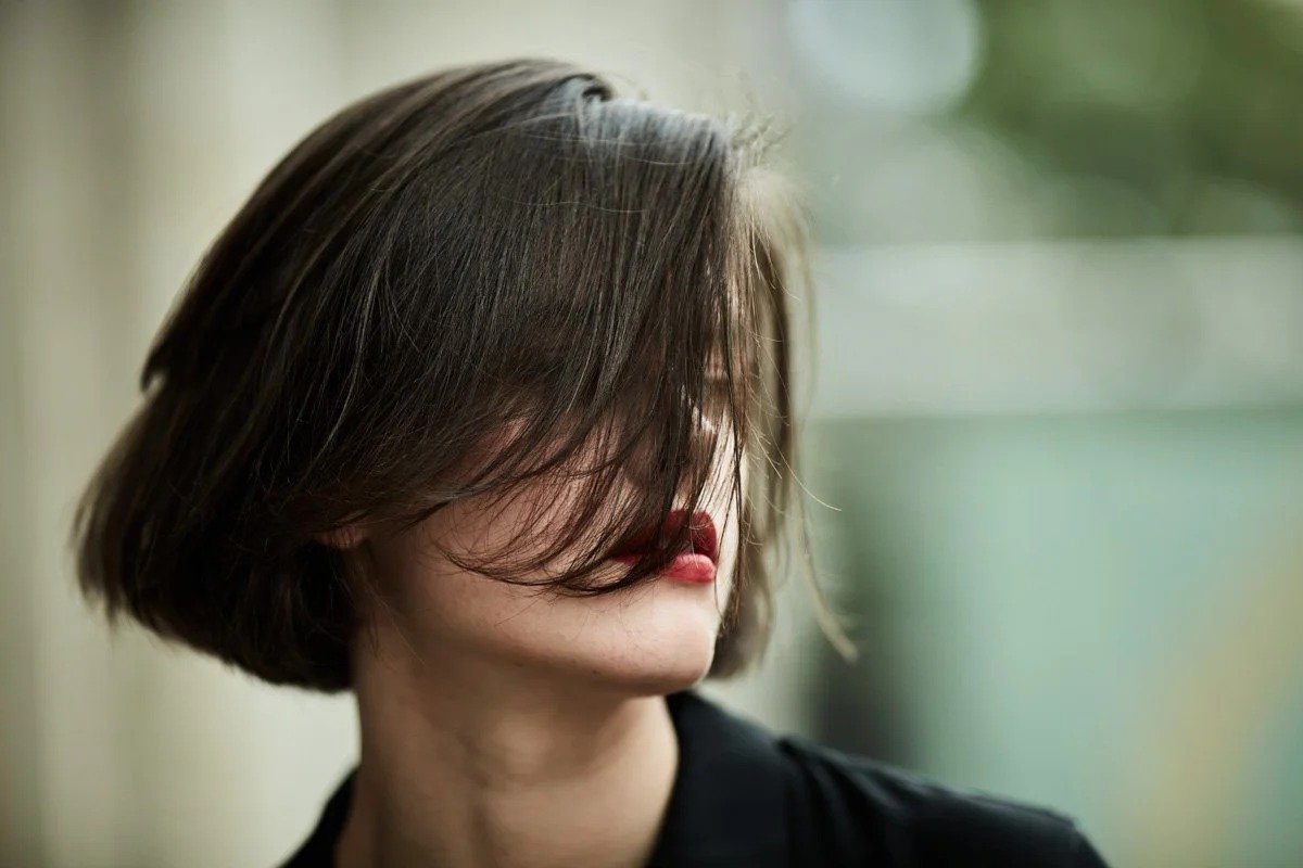 A woman with a short bob haircut covering part of her face with her hair, wearing red lipstick and a black top, with a blurred outdoor background.