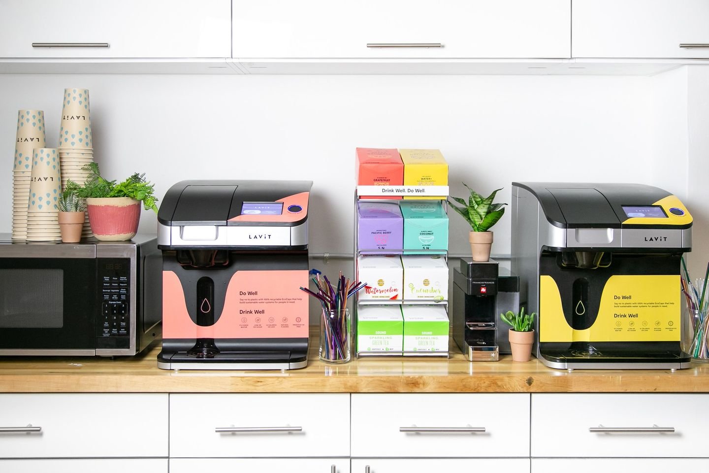 Two Lavit commercial beverage dispensers with different colored panels, a microwave, a small plant, and various boxes of drink mixes on a wooden counter in a cafe or break room.