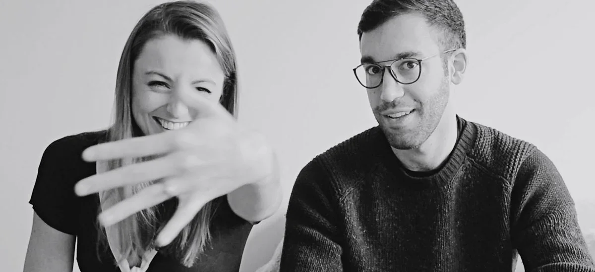 A woman showing her hand towards the camera with a smile, and a man sitting next to her, smiling and wearing glasses, in a black-and-white photo.