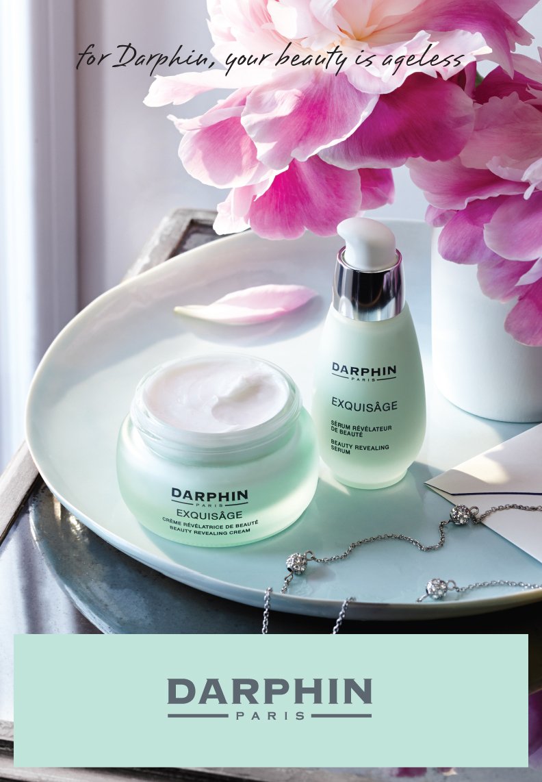 Beauty products on a white tray with pink peony flowers, including Darphin Exquisage beauty revealing cream and serum, near a window with soft natural light, and a silver necklace with clear stones. The image features elegant, feminine skincare items