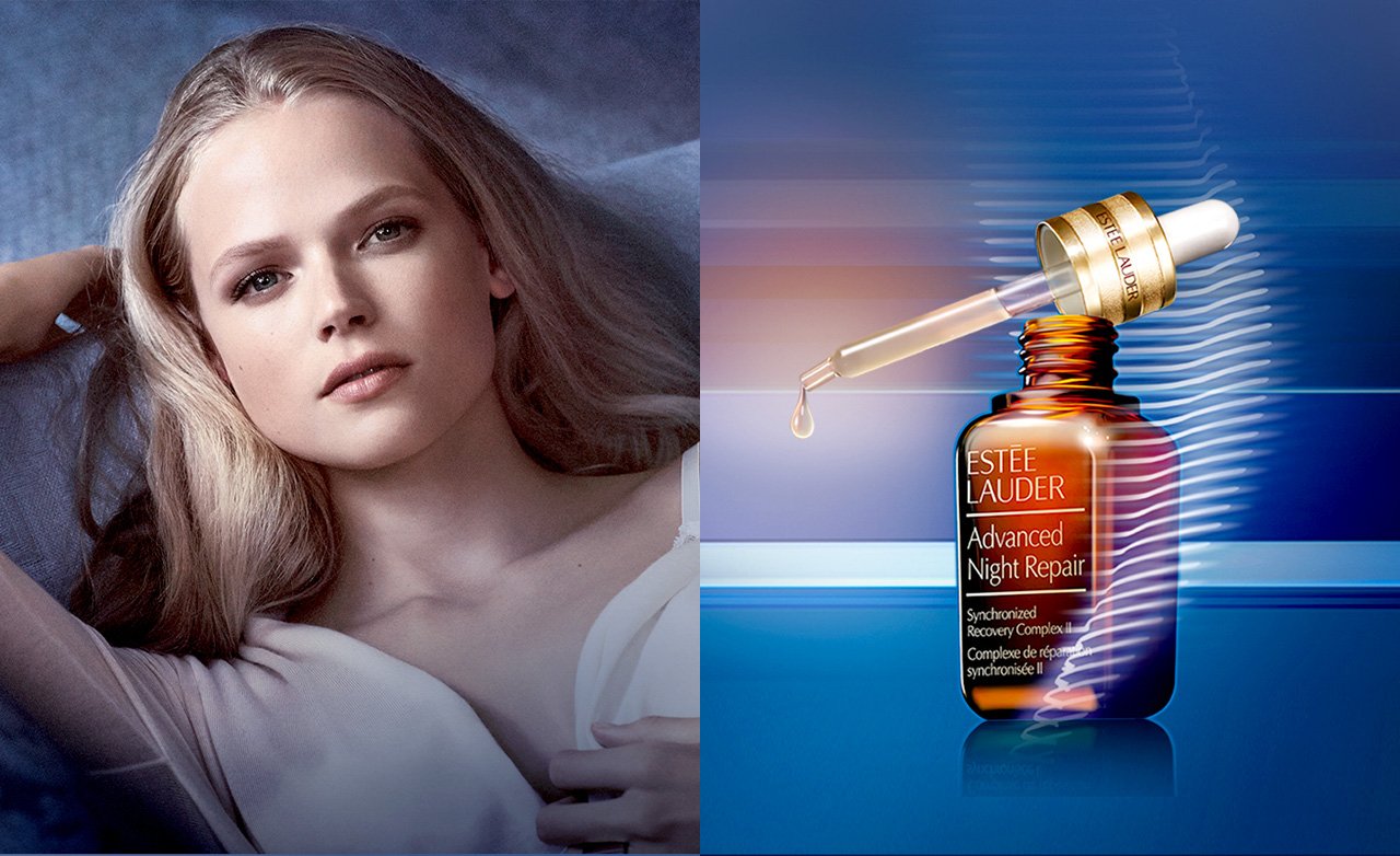 A woman with long blonde hair lying down, looking at the camera, with a bottle of Estee Lauder Advanced Night Repair serum next to her. The serum dropper is releasing a drop into the bottle.