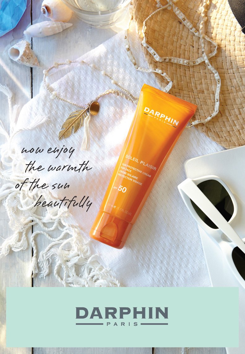 A tube of Darphin Soleil Plaisir sun protective cream SPF 50 placed on white fabric, surrounded by seashells, sunglasses, and a woven straw hat, with a quote overlaying the image that says 'now enjoy the warmth of the sun beautifully'.
